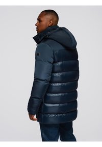Hombre vestido con un abrigo de invierno acolchado azul marino con capucha, de pie en perfil contra un fondo gris liso.