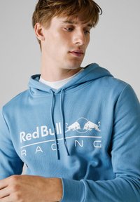 Ljusblå hoodie gjord av mjukt tyg. Har en känguruficka, dragsko på huvan och en stor vit "Red Bull Racing"-logotyp på framsidan.