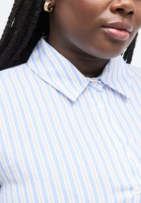 Κοντινό πλάνο ενός ατόμου που φοράει μπλε και άσπρο ριγέ πουκάμισο με κουμπιά και χρυσό σκουλαρίκι σε σχήμα ημισελήνου, με πλεγμένα μαλλιά.