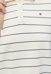 Λευκό πόλο πουκάμισο με λεπτές οριζόντιες μαύρες ρίγες, με τριπλή σειρά κουμπιών και μικρό κόκκινο, λευκό και μπλε λογότυπο στο στήθος.