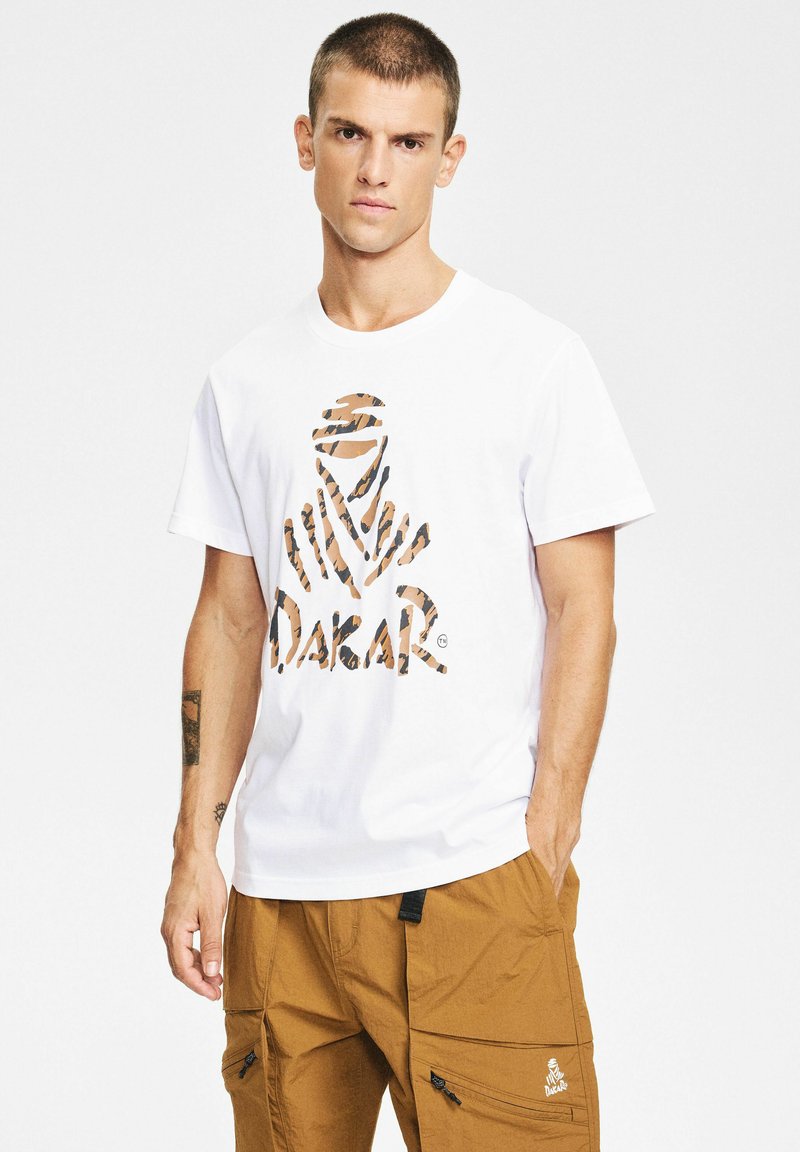 Mężczyzna w białej koszulce z brązowym wzorzystym logo "Dakar" i brązowych spodniach cargo z kieszeniami na zamek, stojący na jednolitym tle.