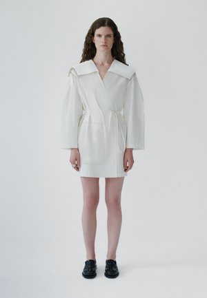 Woman with long curly hair wearing a white belted dress with wide collar and black shoes, standing against a plain white background.