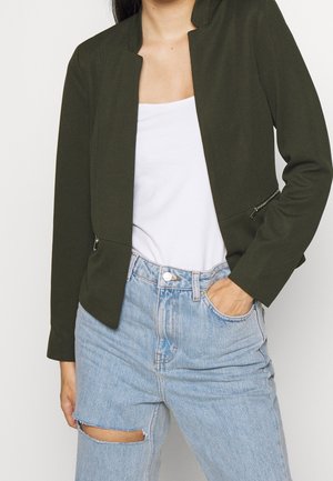Femme portant un blazer vert foncé sur un haut blanc et un jean déchiré bleu clair, avec la main droite dans la poche, devant un fond uni.