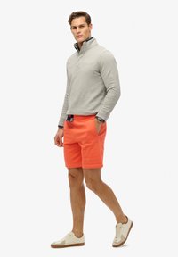 Uomo in piedi con una mano in tasca, indossando un pullover grigio, pantaloni corti arancioni, polsini a quadri, scarpe da ginnastica bianche e un orologio, su uno sfondo bianco.