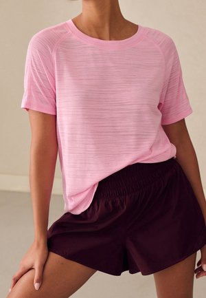 Personne portant une chemise rose clair à manches courtes et un short bordeaux foncé, debout avec une main sur la cuisse.