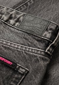 Tela de mezclilla negra con un parche texturizado que presenta letras en relieve. Incluye una pequeña etiqueta rosa cerca de la cintura.