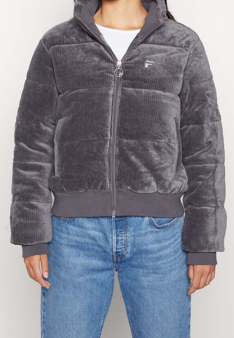 Veste matelassée en velours côtelé gris avec fermeture éclair à l'avant, capuche et poignets côtelés. Présente un logo discret sur la poitrine et une finition épaisse et texturée.