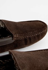 Chaussons en suède brun avec une doublure intérieure douce et texturée, présentant un design cousu et un bout rond. La semelle présente des détails de grip minimal.