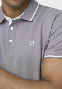 Man in een grijze polo met witte knopen, witte kraagaccenten en een klein wit geometrisch logo op de borst.