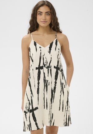 Kobieta ubrana w kremową, bezrękawnikową sukienkę z cienkimi ramiączkami i wyrazistym pionowym wzorem czarnych pociągnięć pędzla, stojąca z rękami w kieszeniach.