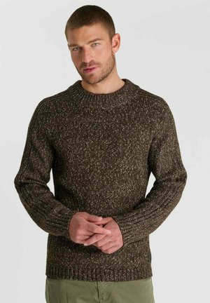 Uomo con capelli corti e barba chiara che indossa un maglione marrone screziato e pantaloni verdi, mani giunte davanti.