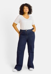 Jean flare bleu foncé avec poches avant et couture dorée, associé à un T-shirt blanc à col en V. Des chaussures décontractées complètent le look.