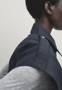 Granatowa kurtka z wyprofilowanym kołnierzem, z metalowymi detalami i teksturowanym materiałem, noszona na szarym długim rękawie.