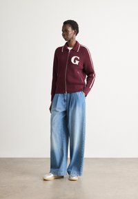 Giacca a zip in maglia bordeaux con texture a coste e righe bianche, contraddistinta da un logo bianco "G". Abbinata a pantaloni di jeans azzurri a gamba larga.