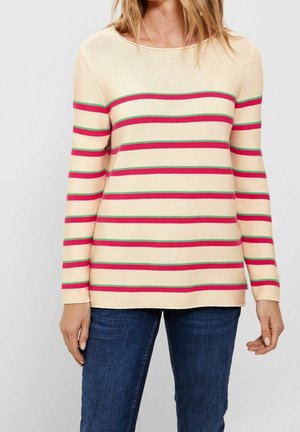 Femme portant un pull crème à manches longues avec des rayures horizontales rouges et vertes, associé à un jean bleu, sur un fond clair uni.