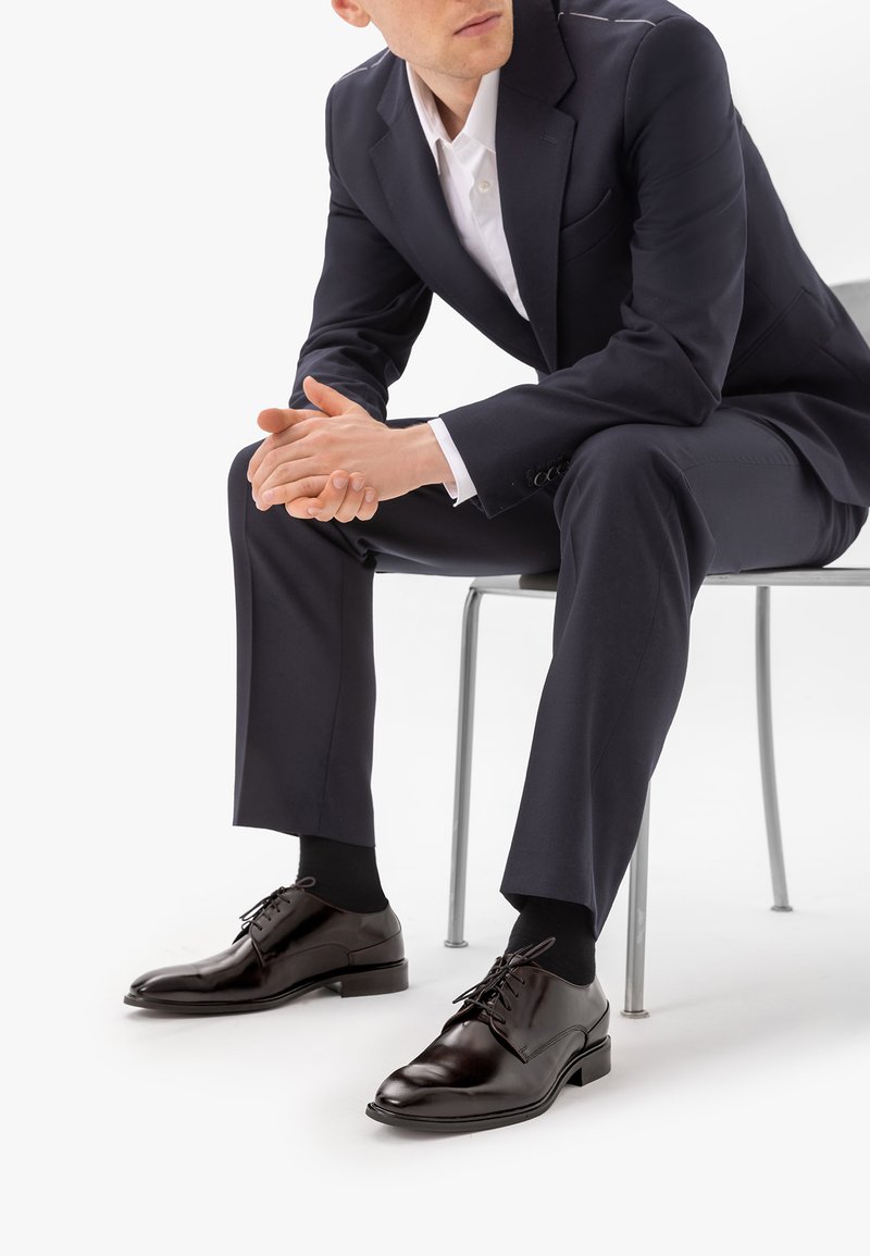 Homme en costume sombre et chemise blanche assis sur une chaise, mains jointes, portant des chaussures habillées en cuir noir poli et des chaussettes noires sur fond blanc.