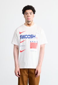 Balts kokvilnas T-krekls ar zilu "SWOOSH" tekstu, sarkaniem akcentiem un režģa rakstu grafiku. Īsas piedurknes un atbrīvota piegriezuma.