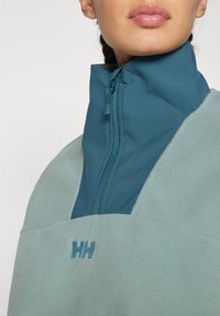 Sudadera de forro polar en un suave verde menta con un alto cuello, media cremallera en azul oscuro y un sutil logo en un color a juego en el pecho.
