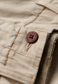 Tessuto di cotone beige con dettagli di cucitura visibili, caratterizzato da un bottone rotondo in plastica marrone con la scritta "SUPERDRY" impressa sulla sua superficie.