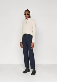 Naine seisab, seljas kreemivärvi fliisist neljandik-zipp-sviiter, tumesinine sirge säärega püksid, mustad tossud ja beežid kassinäoga prillid, taustaks ühtlane valge pind.