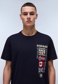 Mørkeblå bomulds t-shirt med vertikal grafisk print med tekst og flag fra Canada, Norge og Storbritannien på venstre side.