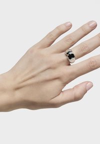 Bague en argent avec une finition polie, ornée d'un design complexe de fleur noire, portée sur une main avec du vernis à ongles neutre.