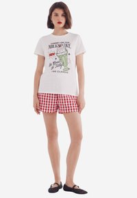 Witte graphic t-shirt met een milkshake-print. Gepaard met rood-wit geruite shorts. Model draagt zwarte platte schoenen.