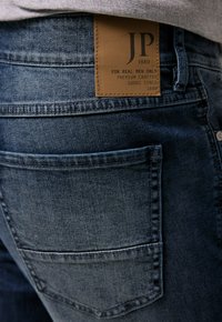 Vaqueros de mezclilla azul con una etiqueta de cuero con texto en relieve, diseño de cinco bolsillos y un ligero desvanecimiento. El material es suave con un corte clásico.