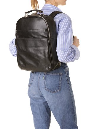 Persoon draagt een lichtblauw gestreept shirt en spijkerbroek, met een stijlvolle zwarte leren rugzak met ritssluitingen op de rug.