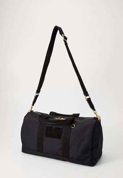 Black duffel bag made of durable fabric, featuring a zippered top, short handles, and a removable shoulder strap with gold hardware accents.
