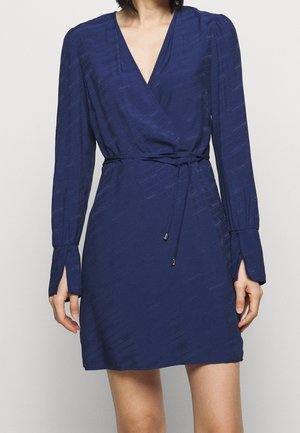 Nainen, jolla on yllään laivastonsininen pitkähihaista kietaisumekkoa V-aukolla ja ohut vyötärölle solmittava vyö.