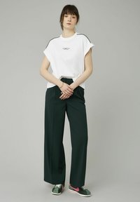 T-shirt en coton blanc à manches courtes, rayures noires sur les épaules et logo. Pantalons larges de couleur vert foncé. Baskets vertes avec des accents roses.