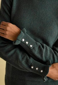 Dunkelgrüner gestrickter Pullover mit gerippten Bündchen und drei metallischen Knöpfen sowie einer strukturierten Oberfläche. Enganliegendes Design aus weichem, warmem Material.