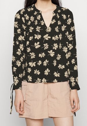 Schwarze Bluse mit langen Ärmeln und weißem Blumenmuster, V-Ausschnitt und Bindebändern an den Handgelenken. Kombiniert mit hellrosa Shorts.