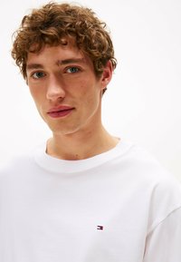 T-shirt bianca in cotone con scollo rotondo. Presenta un piccolo logo ricamato in rosso, bianco e blu sul petto a sinistra. Tessuto liscio.