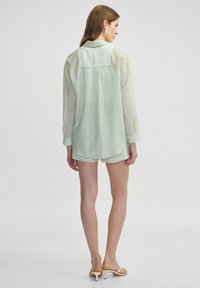 Camisa de manga larga verde claro con un corte relajado y cuello abotonado. Textura semitranslúcida con un brillo sutil. Combinada con pantalones cortos y sandalias.