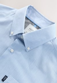 Camicia azzurro chiaro con bottoni, tessuto di cotone, collo classico, bottoni bianchi, taschino sul petto, etichetta con logo sull'orlo, texture liscia.