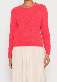 Ljusrosa stickad tröja med rund halsringning, långa ärmar och avslappnad passform, bärs över en plisserad krämfärgad kjol.