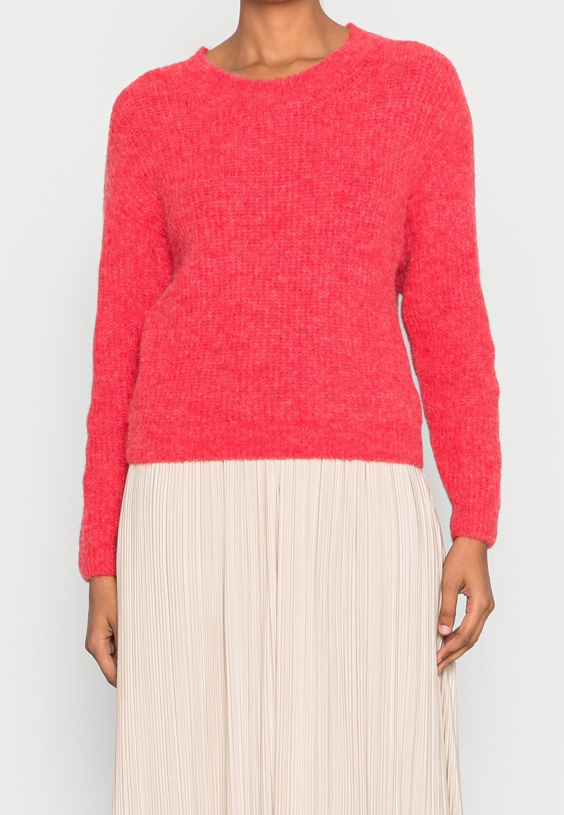 Ljusrosa stickad tröja med rund halsringning, långa ärmar och avslappnad passform, bärs över en plisserad krämfärgad kjol.