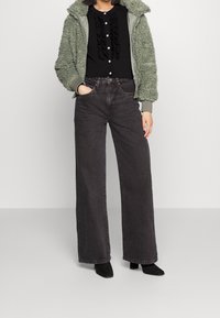 Zelená, nadýchaná zipová bunda přes černou halenku s volány a tmavě šedé široké džíny s vysokým pasem. Černé kotníkové boty dokreslují outfit.