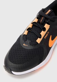 Chaussure de sport noire avec une tige en mailles, des accents orange et une semelle en caoutchouc texturé. Elle dispose de lacets et d'un logo Nike sur le côté.