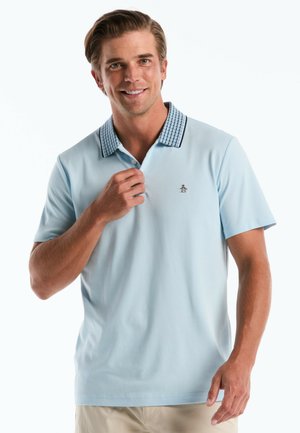 Lächelnder Mann, der ein hellblaues Poloshirt mit gemustertem Kragen und kleinem eingesticktem Logo richtet, trägt beige Hose vor weißem Hintergrund.