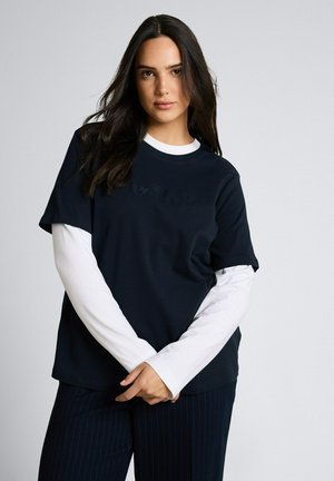 Kobieta o długich ciemnych włosach, ubrana w czarną koszulkę z krótkim rękawem na białej bluzce z długim rękawem, stoi z założonymi rękami na przodzie.