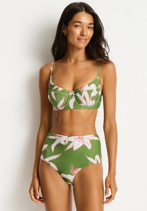 Vrouw draagt groene bloemenprint hoge taille bikini met dunne bandjes en witte bloemenpatronen, staand tegen een effen lichte achtergrond.