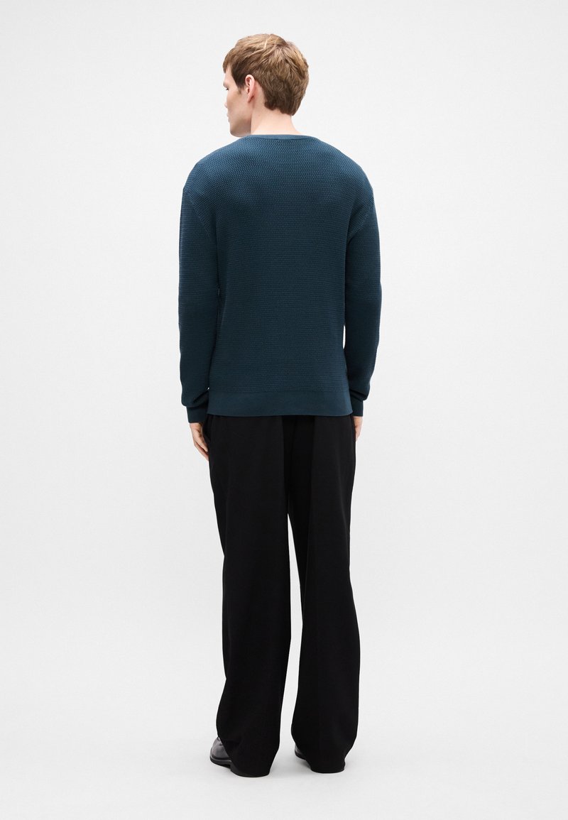 Dark teal knit sweater with a textured pattern and ribbed cuffs, paired with loose black trousers. View from the back, showing the neckline.