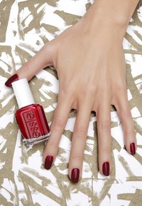 Rode nagellakfles met witte dop naast een hand die verzorgde vingers laat zien. De nagels zijn glanzend en diep rood. De achtergrond heeft gestructureerde, abstracte patronen.
