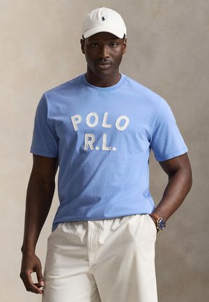 Homme portant un t-shirt Polo R.L. bleu clair, une casquette blanche avec logo, un pantalon blanc et une montre-bracelet, posant sur un fond neutre.