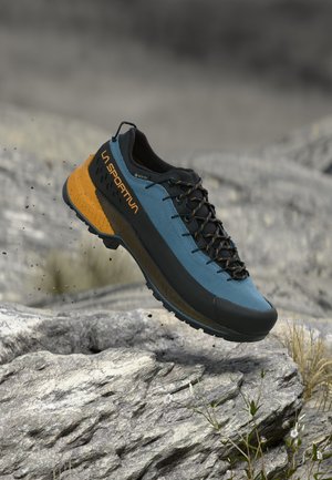 Chaussure de randonnée La Sportiva bleue et orange avec des lacets noirs flottant au-dessus d'un terrain rocheux avec de petites plantes.