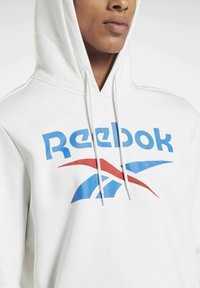 Hvit hettegenser med et frontgrafikk som viser Reebok-logoen i blått og rødt. Justerbare snorer er festet til hetten.