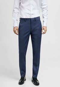 Marineblaue, taillierte Hose mit schmalem Schnitt, aus einem glatten Stoff, mit Gürtelschlaufen und einem Bügelfalte auf der Vorderseite, kombiniert mit schwarzen Schuhen.